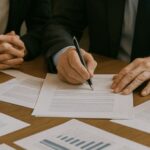 Quatro empresários em trajes formais participam de uma reunião em uma sala iluminada, analisando e assinando documentos sobre a mesa
