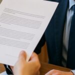 pessoa entrega um contrato a um funcionário de cartório sentado à mesa, em ambiente interno iluminado naturalmente, com expressão atenta e realismo nos detalhes