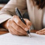 Como é feita a escritura publica de cessão de direito? 1 Pessoa adulta assinando um documento em um cartório, em ambiente interno institucional, com iluminação suave e natural