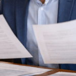 Funcionário de cartório sentado à mesa, comparando atentamente dois documentos em um ambiente institucional organizado