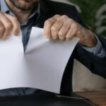 Contrato de compra e venda de imóvel pode ser cancelado? 3 Pessoa rasgando um contrato em um ambiente de escritório, com expressão séria e uma maquete de imóvel sobre a mesa, representando dúvidas ou cancelamento em uma negociação imobiliária