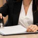 Funcionária de cartório registrando um documento com carimbo sobre o balcão, em ambiente moderno e profissional, enquanto atende uma pessoa ao seu lado